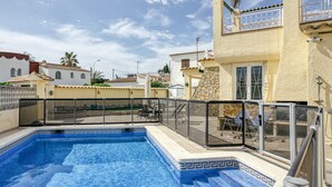 Piscina con borde de azulejos, un porche cubierto con sillas y vistas a casas vecinas.