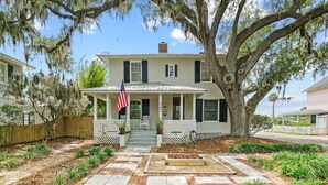 A two-story house with a porch, an American flag, and a large tree in the yard. - The Charlotte House - Pool, Hot tub, Night of Lights! (Downtown St. Augustine)