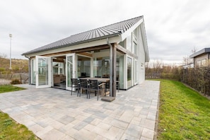 A modern house with a patio, a dining area with chairs and a table, and a large glass door leading to the interior.