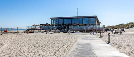 A modern beachside building with a glass facade, a concrete walkway leading to it, and beach umbrellas in the background.