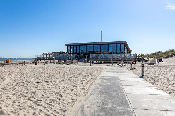 A modern beachside building with a glass facade, a concrete walkway leading to it, and beach umbrellas in the background.