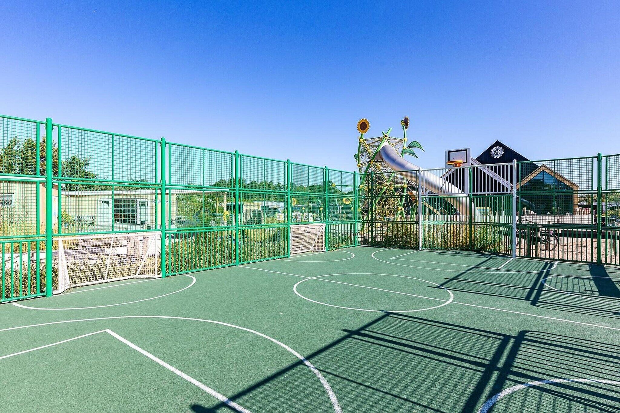 En udendørs basketballbane med et grønt hegn, en basketballkurv og en bygning i baggrunden.