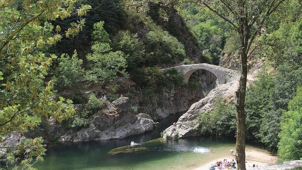 A stone bridge over a river, surrounded by trees and a sandy beach with people.