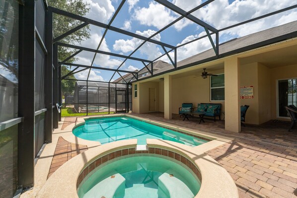 A covered outdoor swimming pool with a hot tub, surrounded by patio furniture and a glass enclosure.