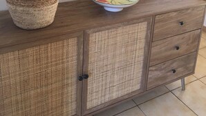 A wooden cabinet with a woven basket containing a plant, a decorative bowl, and a mirror reflecting a dining area with a sofa and cushions.