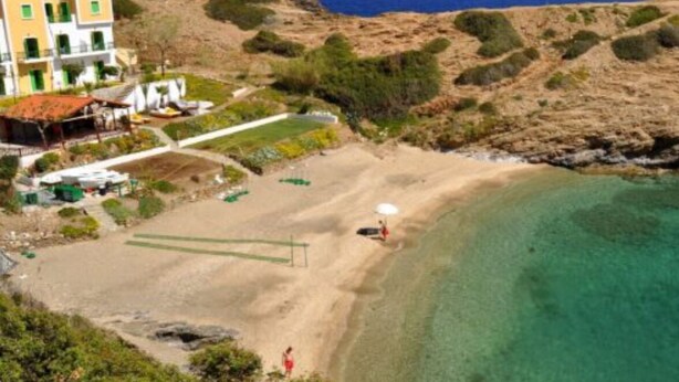 A coastal property with a sandy beach, a clear blue sea, and a hillside with vegetation.