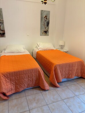 Quarto de hotel com duas camas, um quadro na parede, uma mesa de cabeceira com luminária e piso ladrilhado.