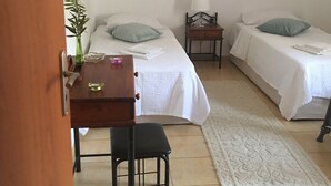 A room with two beds, a small table with a chair, a lamp, and a decorative plant.