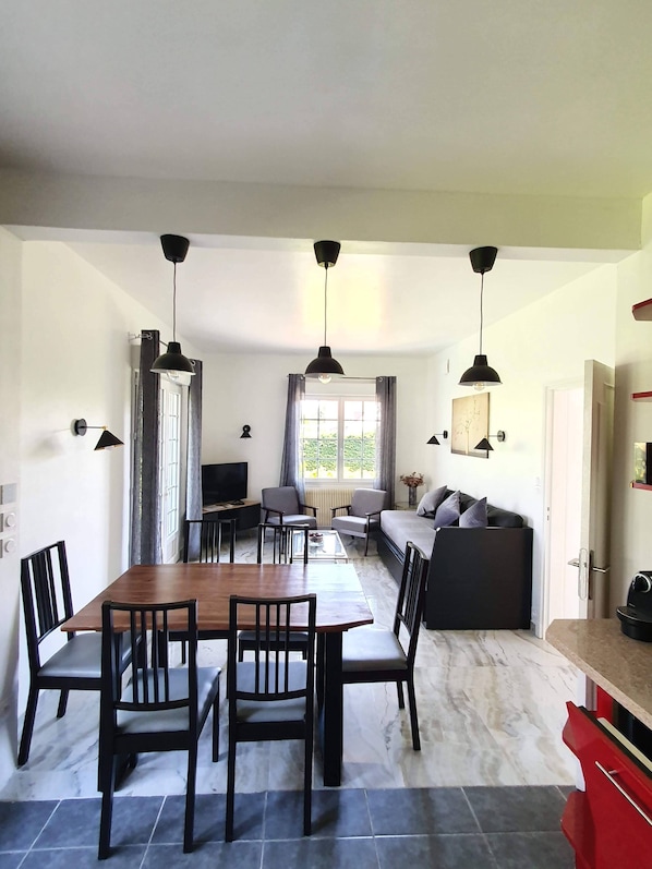A modern living room with a wooden dining table, black chairs, a grey sofa, and a red kitchen counter.