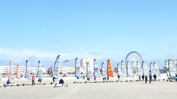 Une plage où l’on peut voir de nombreux voiliers alignés, une grande roue en arrière-plan et des bâtiments le long de la côte.