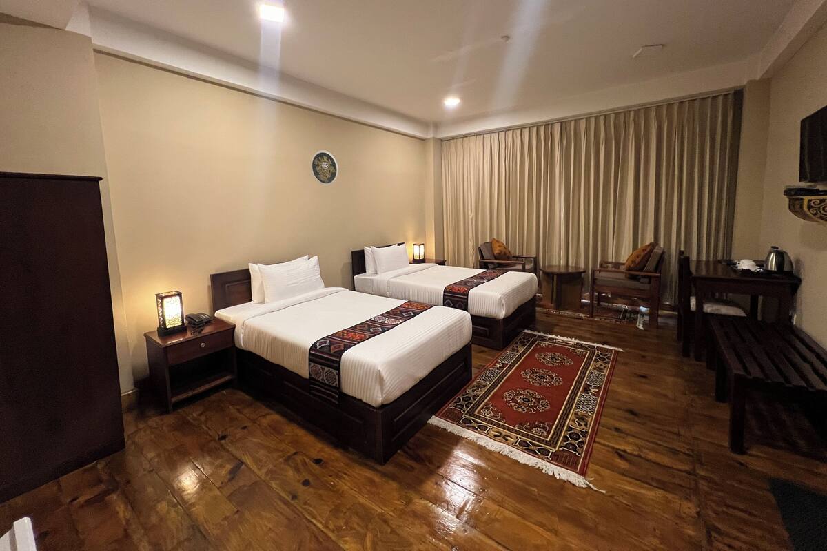 A hotel room with two beds, a desk, a chair, a small table, a clock on the wall, and a window with curtains.