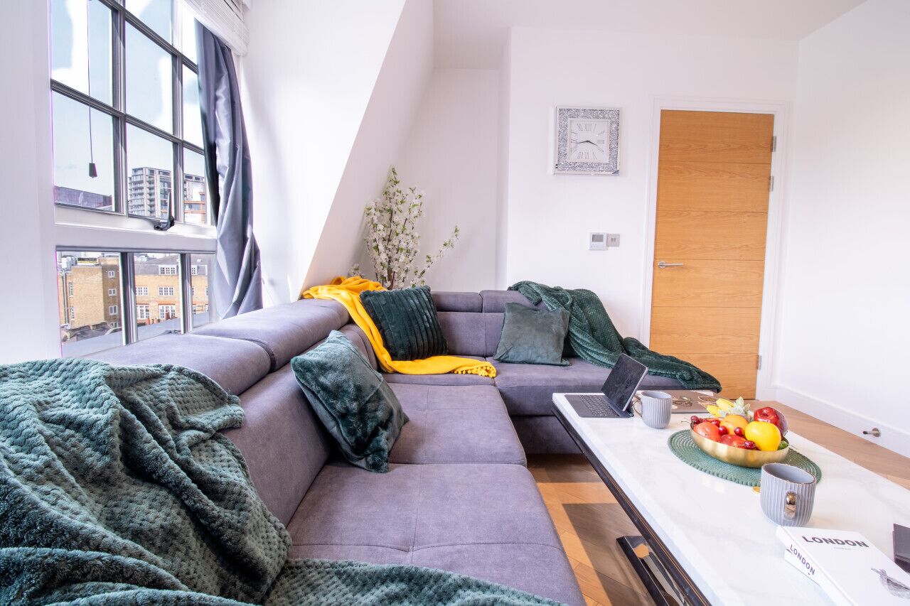 A modern living room with a sectional sofa, a dining table with a laptop, a bowl of fruit, and a wooden door.