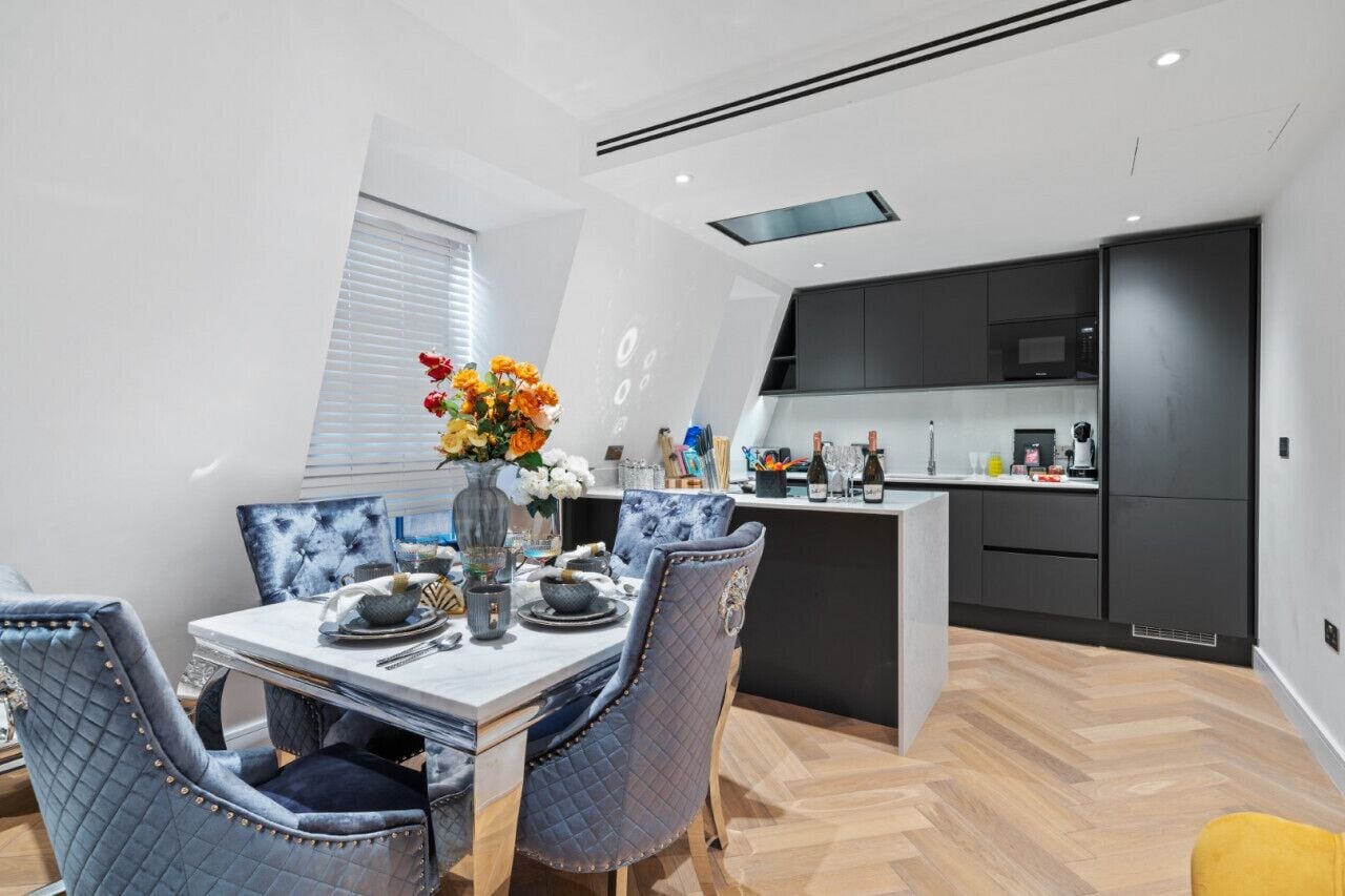 Une cuisine moderne avec des meubles de couleur sombre, un îlot central et un coin repas comprenant une table en marbre et des chaises recouvertes d’un tissu bleu.