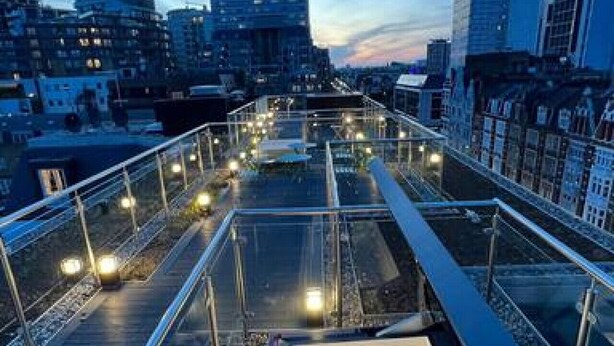 Een dakterras met glazen hekwerk, een trap en een stadsgezicht in de schemering.