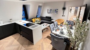 Een moderne keuken met een inbouw eiland, een eetgedeelte met een wit gedekte tafel, en een woongedeelte met een zwarte bank en een decoratieve wand.