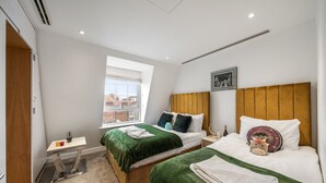 Quarto de hotel com duas camas, uma mesinha, janela com vista e um quadro na parede.
