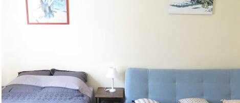 Un dormitorio con cama, mesita de noche con lámpara, sofá y dos cuadros colgados en la pared.
