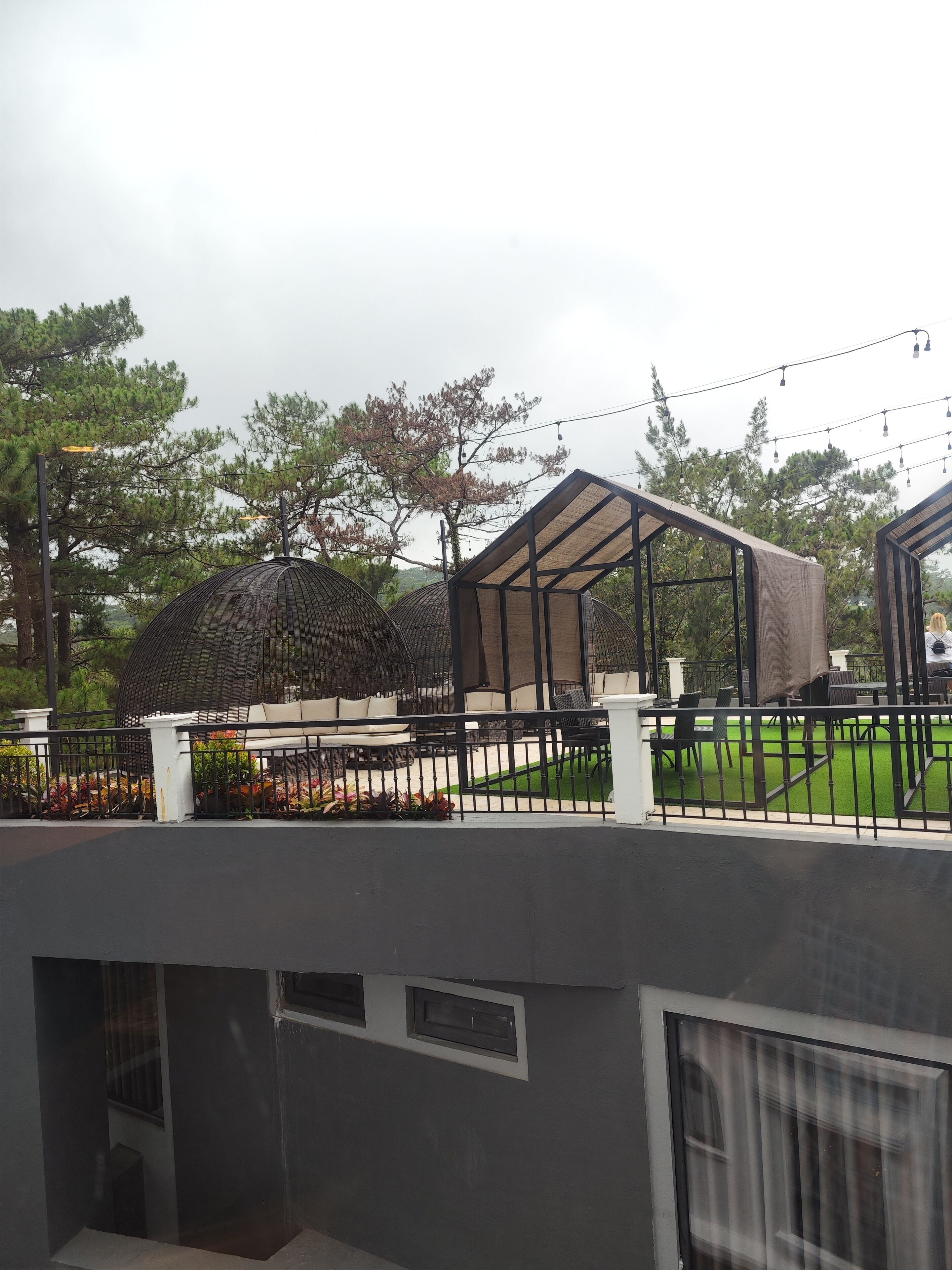A covered outdoor seating area with tables and chairs, surrounded by a fence and greenery.