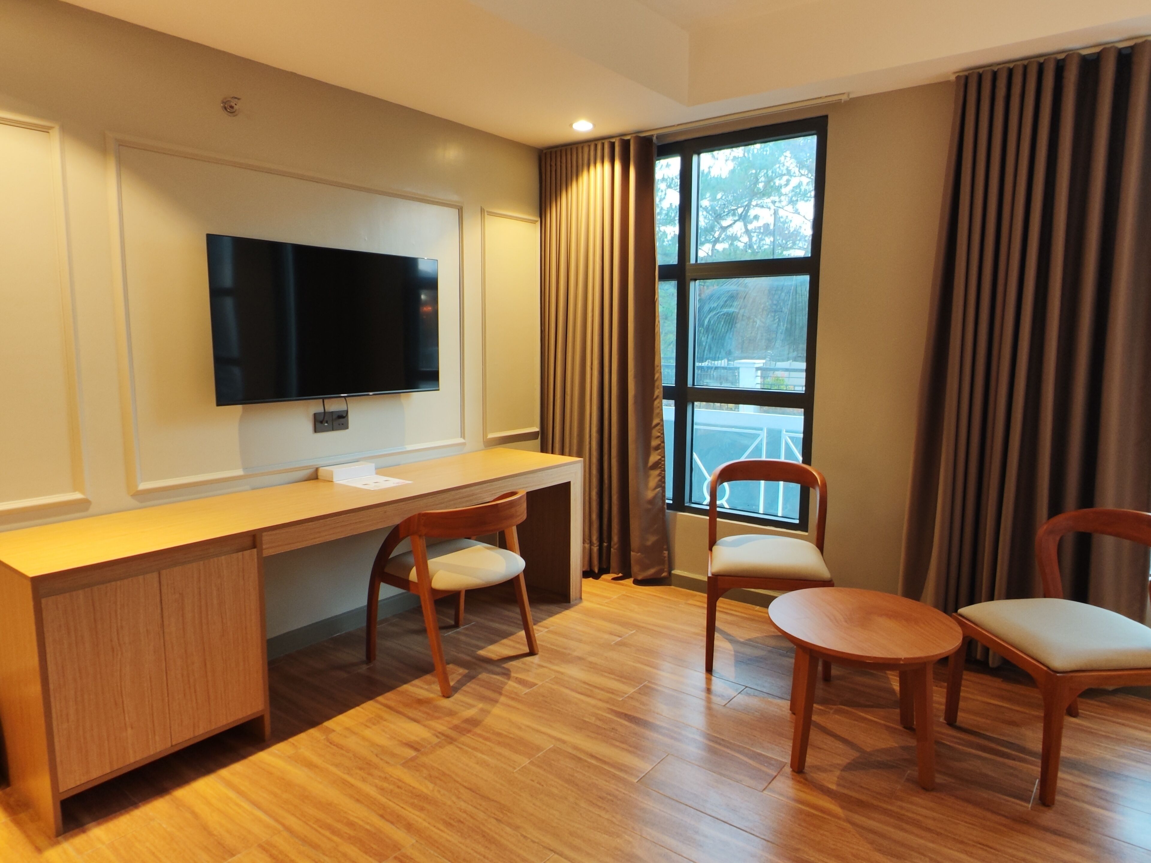 A modern hotel room with a wooden desk, two chairs, a flat-screen TV mounted on the wall, and a large window with curtains.