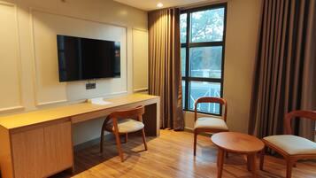 A modern hotel room with a wooden desk, two chairs, a flat-screen TV mounted on the wall, and a large window with curtains.