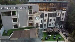 Un hotel llamado HOTEL GRANDE con una destacada torre del reloj, un área de estar al aire libre y un edificio moderno al fondo.