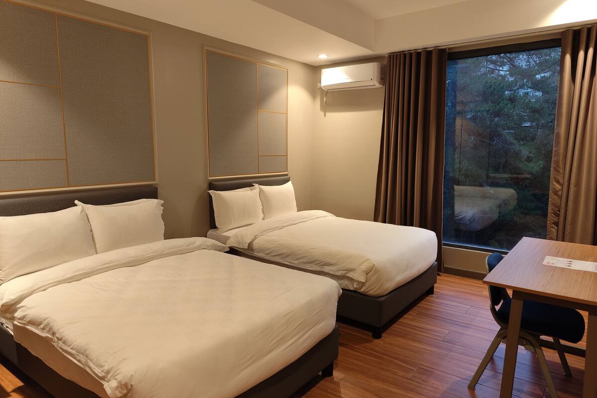 A hotel room with two beds, a wooden floor, a table, and a window with curtains.