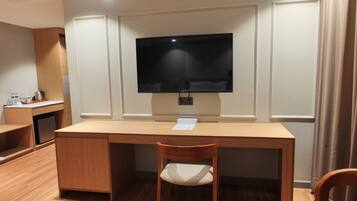 A hotel room with a wooden desk, a flat-screen TV mounted on the wall, a chair, and a small kitchenette area.