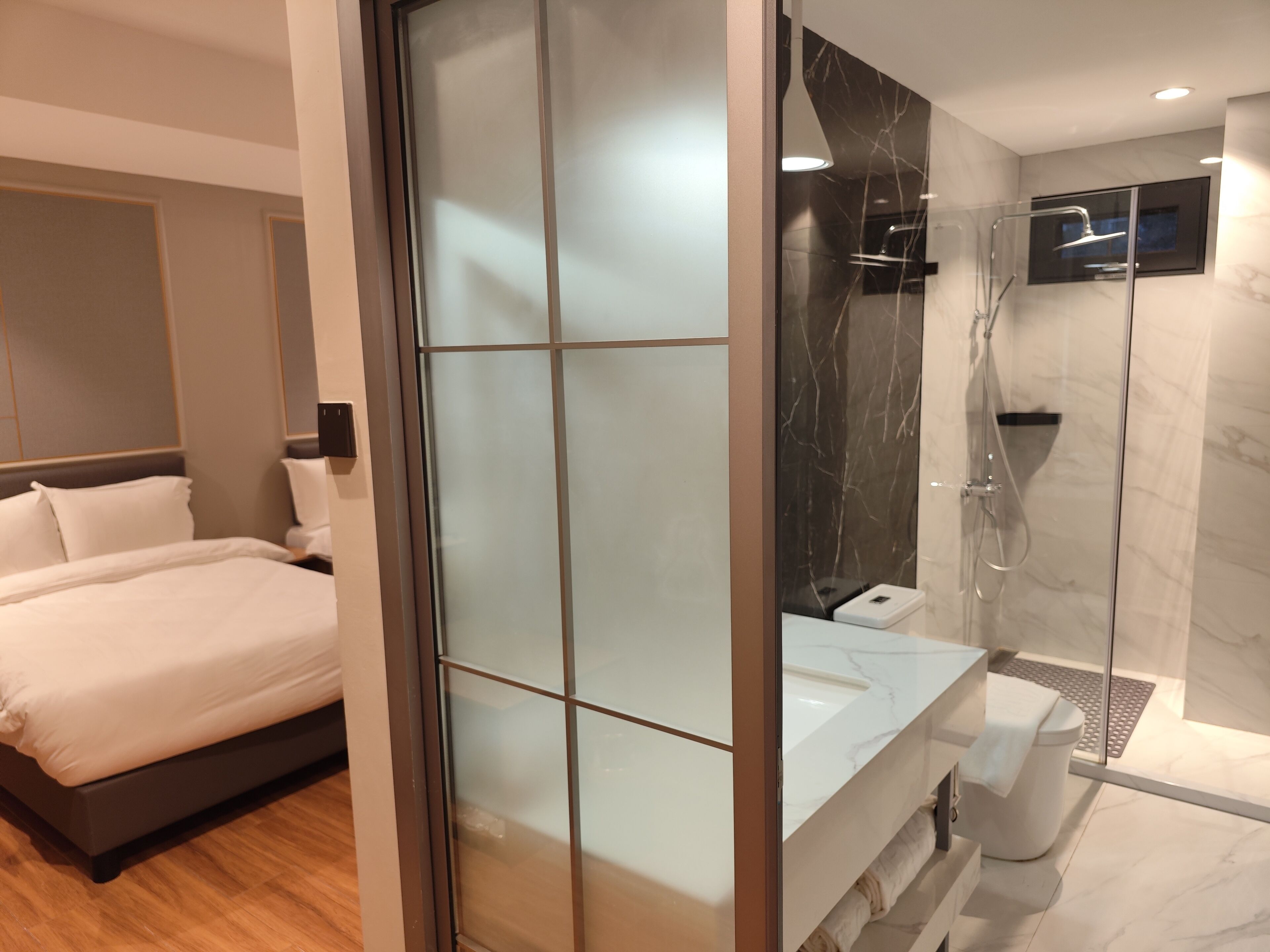 A modern hotel room with a bed, a glass partition, a shower area, and a marble bathroom.