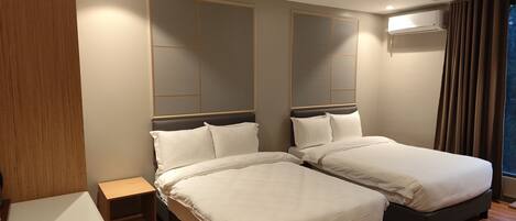 A hotel room with two beds, a wooden floor, a small bedside table, and a window with curtains.