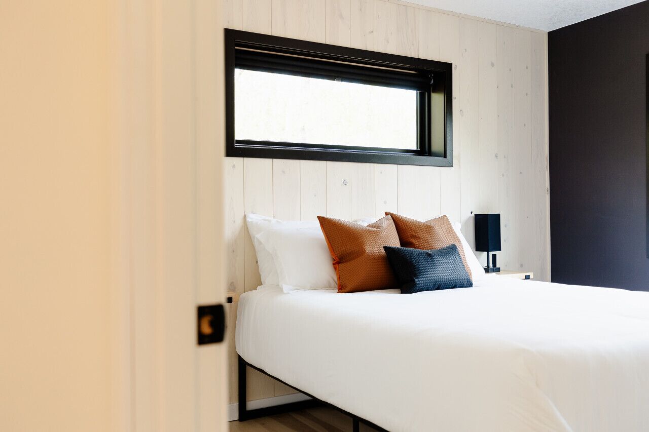 A neatly made bed with white and brown pillows, a black nightstand with a lamp, and a window above the bed.