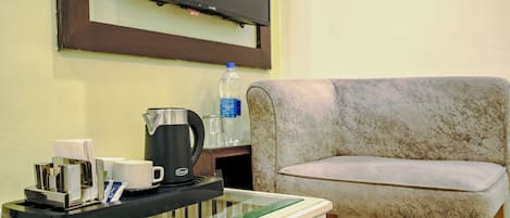 A room with a glass-top table holding a coffee maker, cups, and a bottle of water. A television is mounted on the wall above the table.