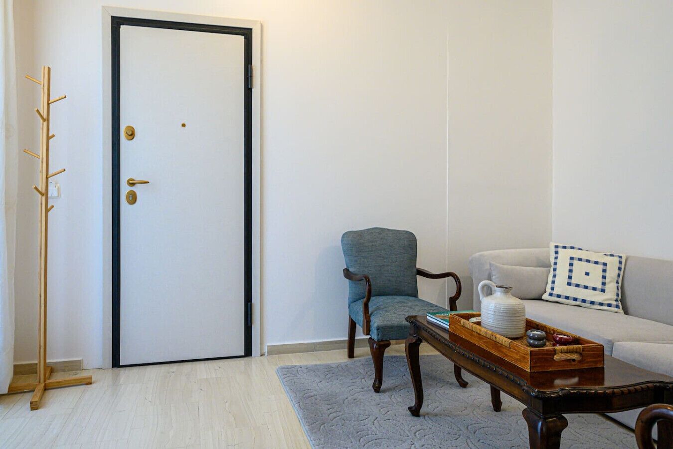 A room with a wooden coat rack, a door with a black frame, a blue armchair, a white sofa with a patterned pillow, and a wooden coffee table with a tray of items.