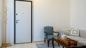 A room with a wooden coat rack, a door with a black frame, a blue armchair, a white sofa with a patterned pillow, and a wooden coffee table with a tray of items. - Anna’s apartment Historic Center (Kalamata)