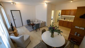 A modern living room with a sofa, chairs, a coffee table, and a dining table. There is a kitchen area with wooden cabinets and a sink. - Anna’s apartment Historic Center (Kalamata)