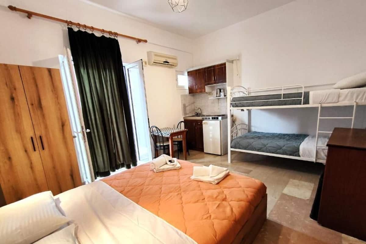 A compact hotel room with a bed, a kitchenette, a wooden wardrobe, and a bathroom.