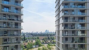 Un immeuble résidentiel de grande hauteur, doté de nombreux balcons, avec une vue sur le paysage urbain en arrière-plan et un parc au premier plan.
