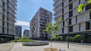 Un immeuble d’appartements moderne situé en milieu urbain, avec de nombreux étages, des balcons et une cour intérieure agrémentée d’un petit arbre et d’une fontaine circulaire.