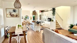 Een moderne keuken met witte kasten, een eethoek met een houten tafel en witte stoelen, en een trap die naar een slaapkamer op een entresol leidt.