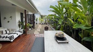 Een terras met een houten dek, een witte tafel met een plant in pot, en een zithoek met kussens.