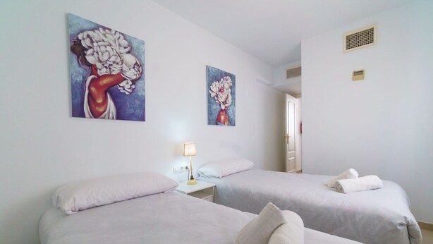 Une chambre d’hôtel avec deux lits, du linge de lit blanc, une table de chevet avec une lampe et deux tableaux aux motifs floraux accrochés au mur.