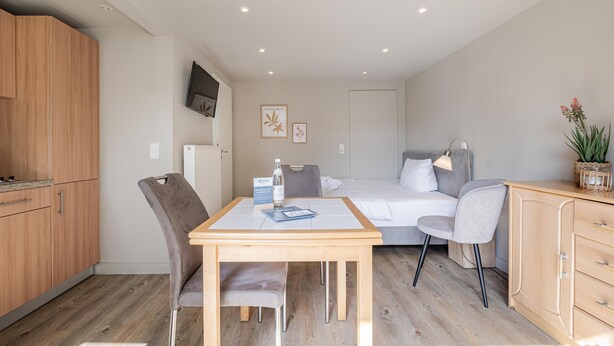 Une chambre d’hôtel compacte avec un coin cuisine en bois, un lit avec une literie grise, une petite table avec une chaise et une lampe de chevet.