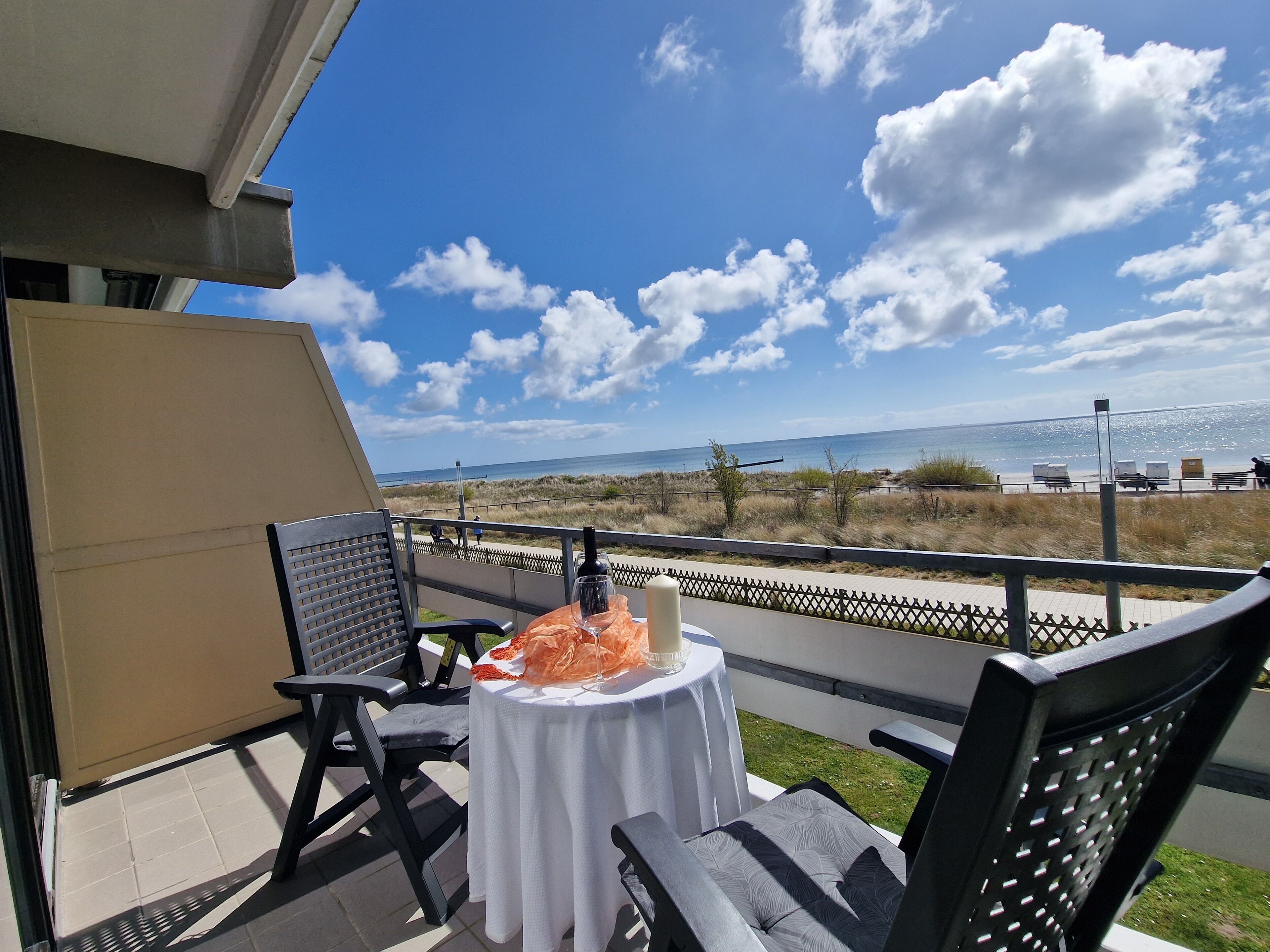 Ein Balkon mit Tisch für zwei Personen, mit Blick auf einen Strand bei blauem Himmel und Wolken.