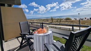 Ein Balkon mit Tisch für zwei Personen, mit Blick auf einen Strand bei blauem Himmel und Wolken.