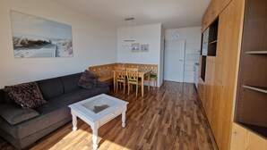Ein gemütlicher Wohnbereich mit einer Couch, einem Esstisch mit Stühlen, einem kleinen weißen Couchtisch, Holzböden, einem Bild an der Wand und einer Tür zu einer Küchenzeile.