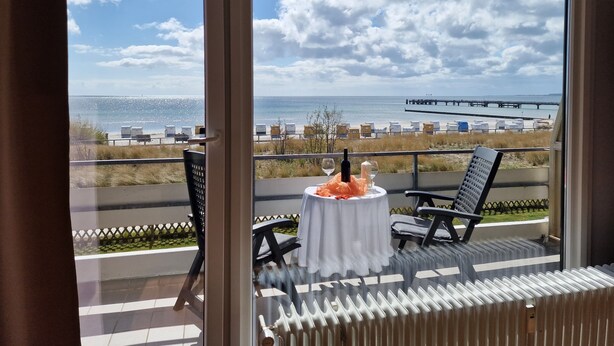 Ein Balkon mit Tisch für zwei Personen, Blick auf einen Strand mit Liegestühlen und einem Steg.