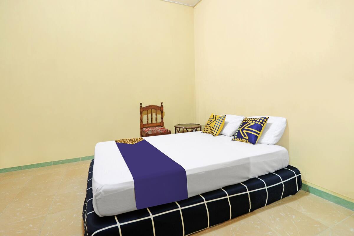 Ein Bett mit weißer Matratze, einer blauen Decke und gemusterten Kissen. Neben dem Bett steht ein Holzstuhl und ein kleiner runder Tisch.
