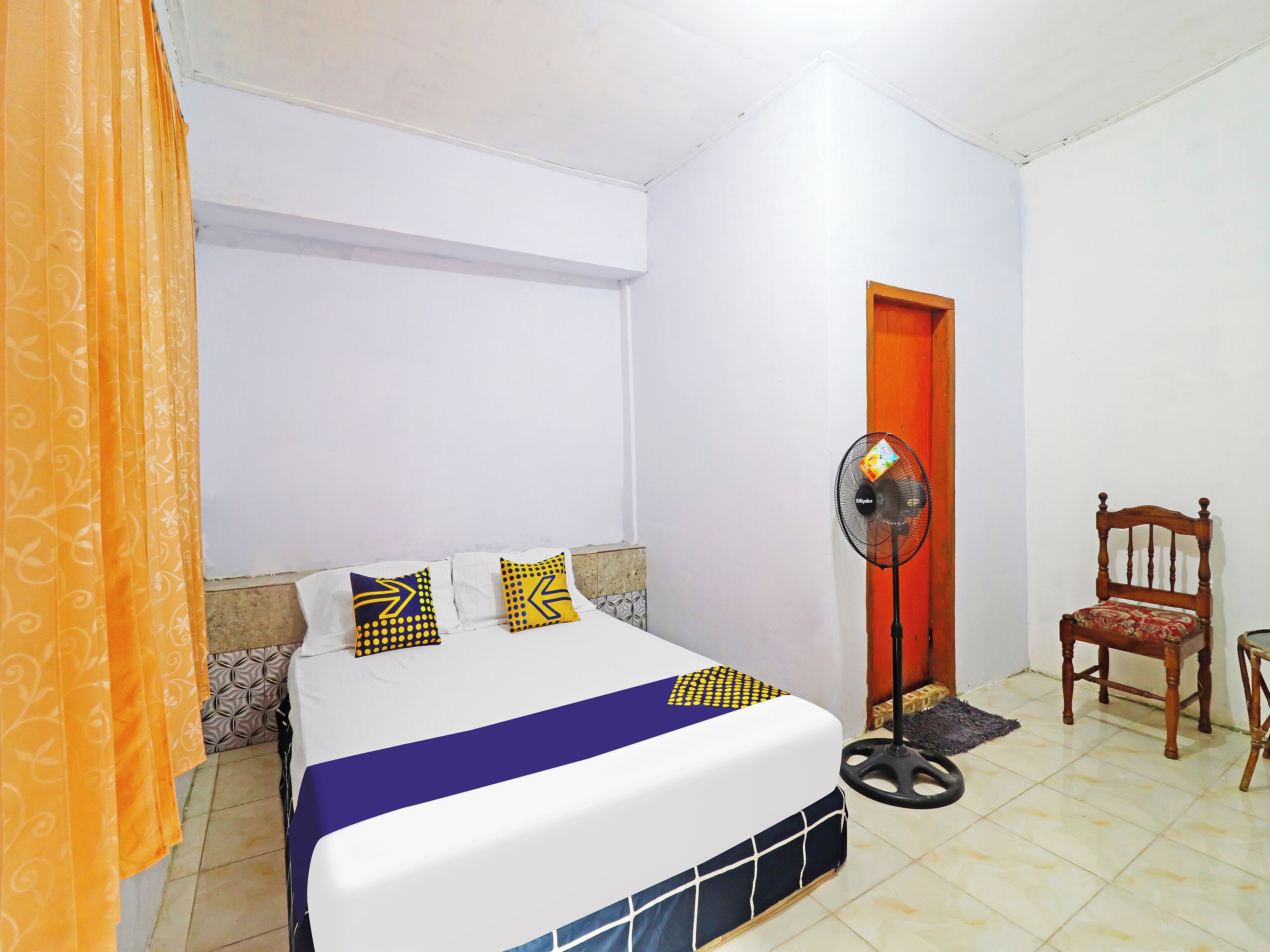 A bedroom with a bed, a standing fan, a wooden chair, and a door.