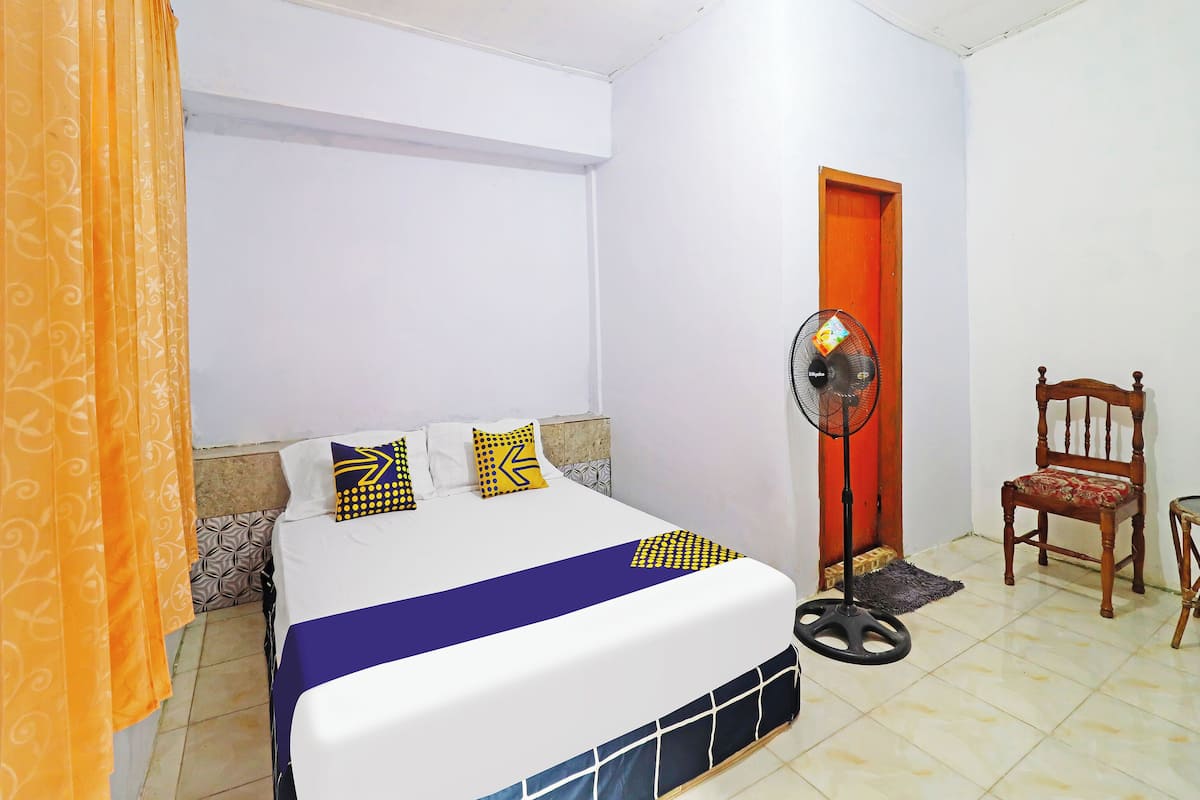 A bedroom with a bed, a standing fan, a wooden chair, and a door.