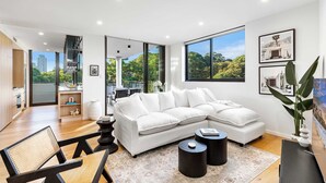 Uma sala de estar moderna com um sofá seccional branco, uma área de refeições com mesa de madeira, e janelas amplas com vista para áreas verdes e edifícios.