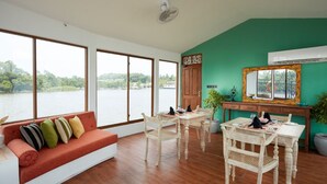Una habitación con piso de madera, un sofá con cojines, una mesa de comedor con sillas, un espejo y vista a una masa de agua a través de amplias ventanas.
